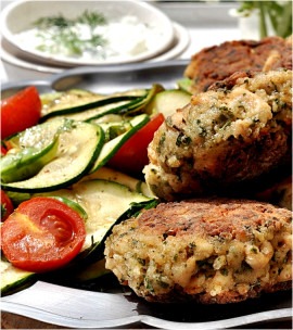 Dwa kotlety warzywne z widocznymi ziołami, podane z grillowaną cukinią, pomidorkami koktajlowymi i sosem tzatziki w tle.