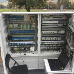 Otwarta szafa sterownicza z rozbudowanym okablowaniem, widoczny laptop na stole obok, krzesło i otoczenie zewnętrzne sugerujące montaż na zewnątrz.