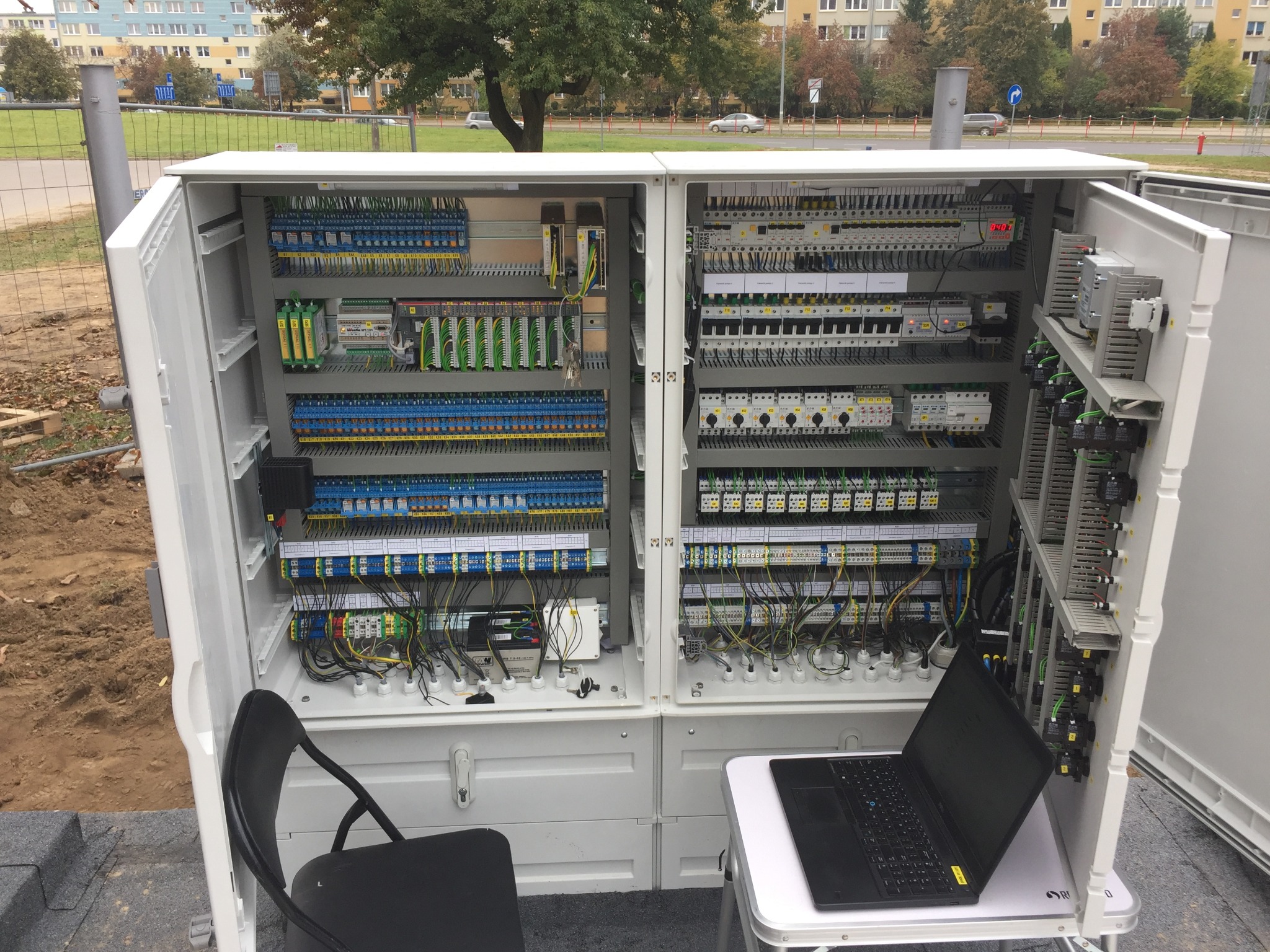 Otwarta szafa sterownicza z rozbudowanym okablowaniem, widoczny laptop na stole obok, krzesło i otoczenie zewnętrzne sugerujące montaż na zewnątrz.