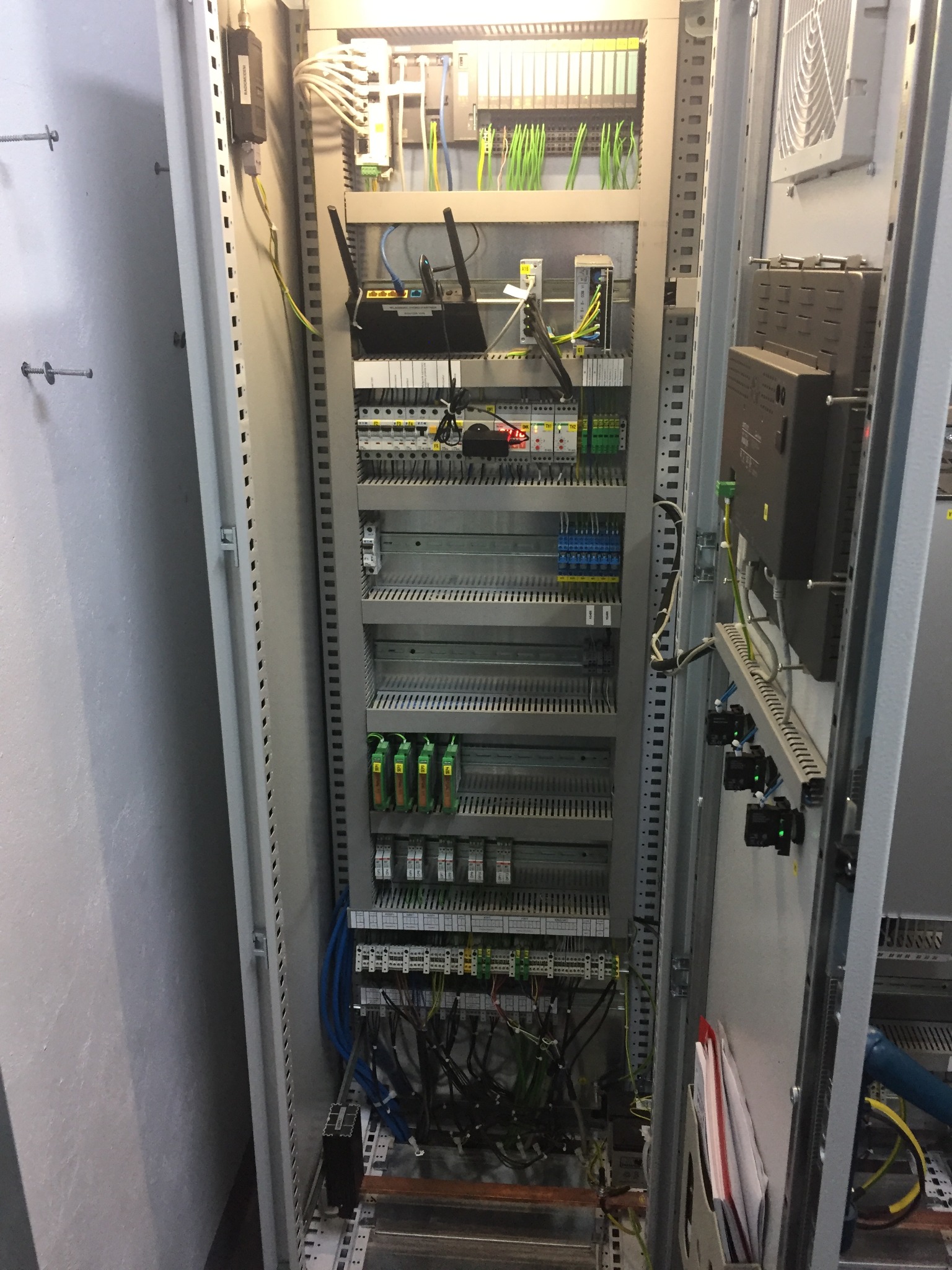 Otwarta szafa sterownicza z widocznymi modułami PLC, okablowaniem, routerem WiFi na górze i elementami automatyki przemysłowej.