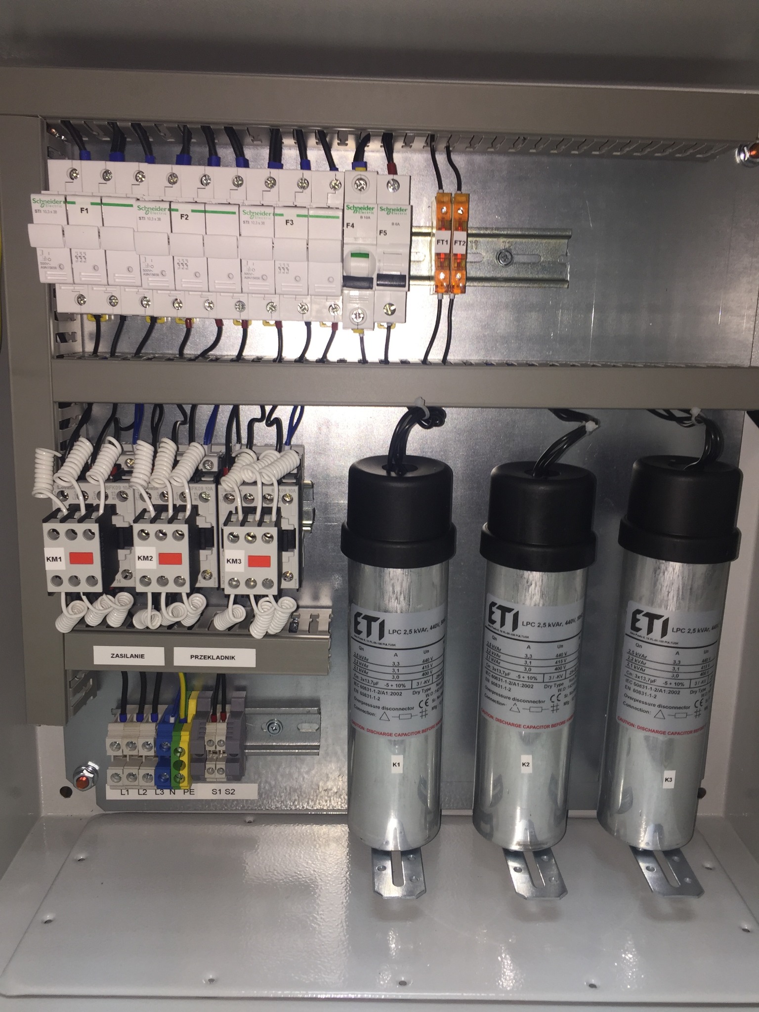 Wnętrze szafy sterowniczej z kondensatorami kompensacyjnymi ETI, stycznikami Levato i zabezpieczeniami Schneider Electric, widoczne okablowanie i oznaczenia.