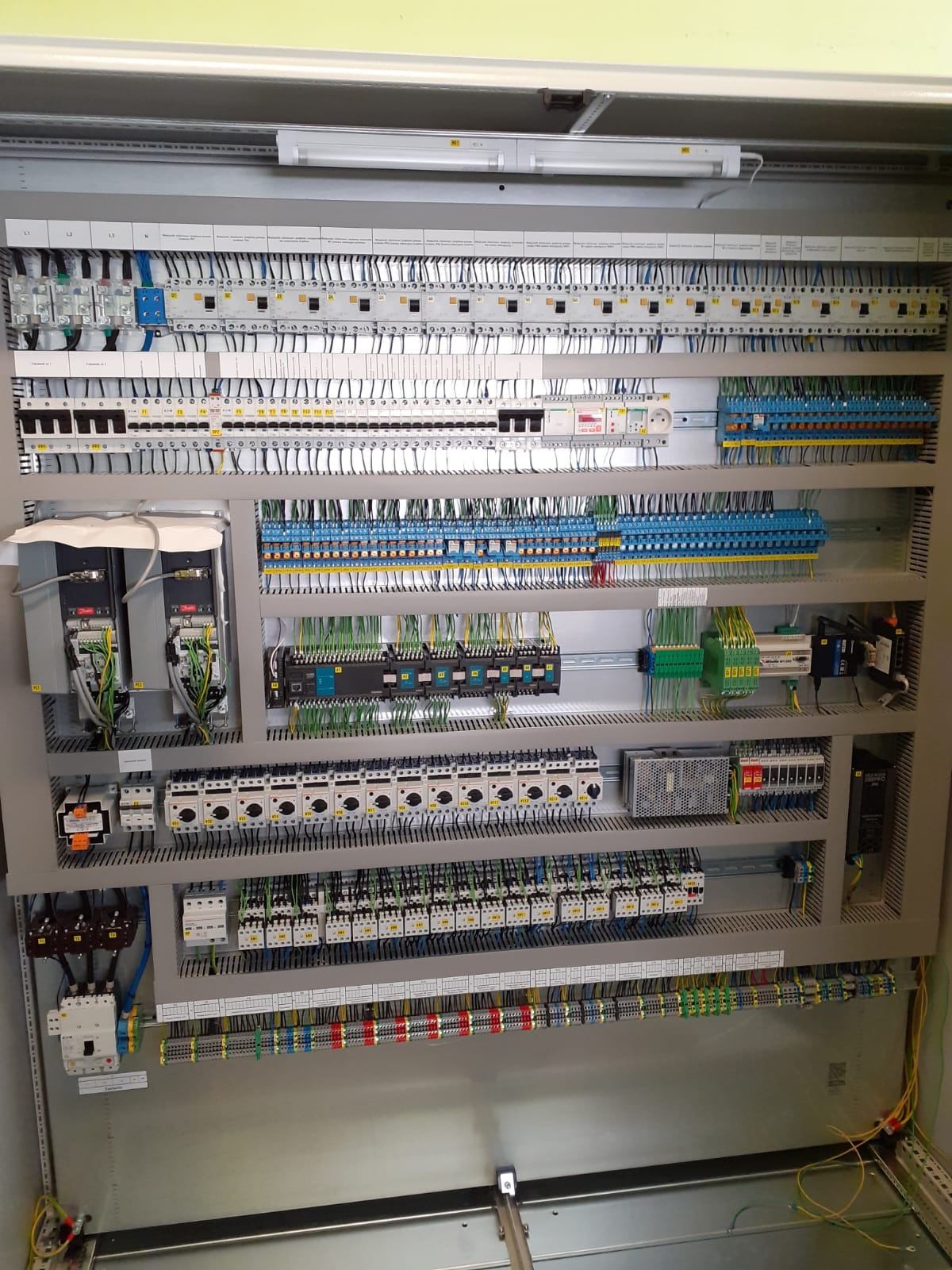 Rozbudowana szafa sterownicza z licznymi przewodami, modułami sterującymi i zabezpieczeniami, starannie okablowana w szarym metalowym panelu, oświetlona lampą jarzeniową.