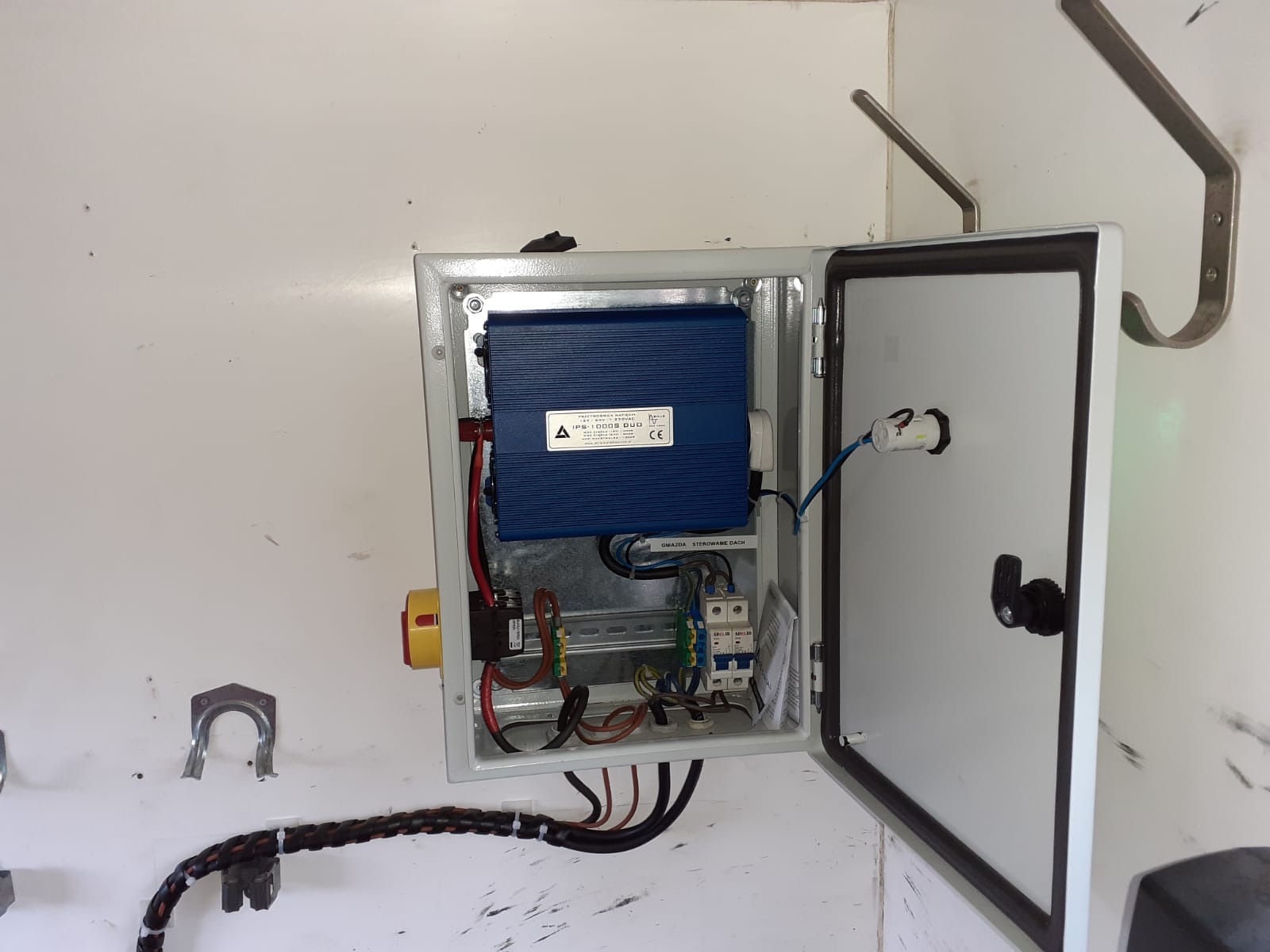 Otwarta skrzynka elektryczna z niebieskim modułem sterującym IPS-1000S DUO, widoczne przewody, bezpieczniki i wyłącznik awaryjny na białej ścianie.