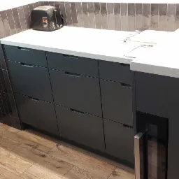 Nowoczesna kuchnia z szarymi szafkami, jasnym blatem i ciemnymi, pionowymi płytkami na ścianie, uzupełniona drewnopodobną podłogą i czarnym tosterem.