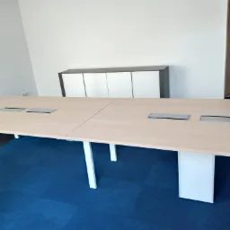 Duży, jasny stół konferencyjny z wbudowanymi gniazdami elektrycznymi i niebieską wykładziną dywanową w tle minimalistycznego biura.