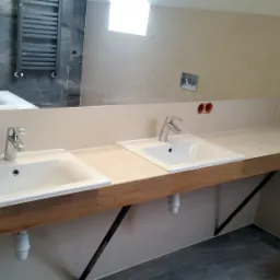 Nowoczesna łazienka z dwiema umywalkami nablatowymi na drewnianym blacie wspartym na metalowych wspornikach, w tle widoczne lustro odbijające wannę i grzejnik drabinkowy.