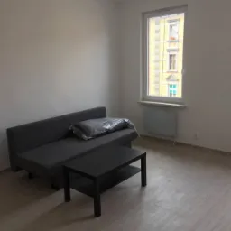 Jasne wnętrze pokoju z szarą sofą, czarnym stolikiem kawowym i podłogą imitującą drewno, widok na budynek z żółtą elewacją przez okno.