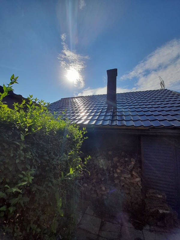 Błyszczący dach z czarnej dachówki ceramicznej w promieniach słońca, widoczny komin i krzew z przodu, ułożone drewno na opał.