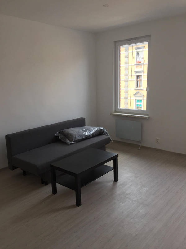 Jasne wnętrze pokoju z szarą sofą, czarnym stolikiem kawowym i podłogą imitującą drewno, widok na budynek z żółtą elewacją przez okno.