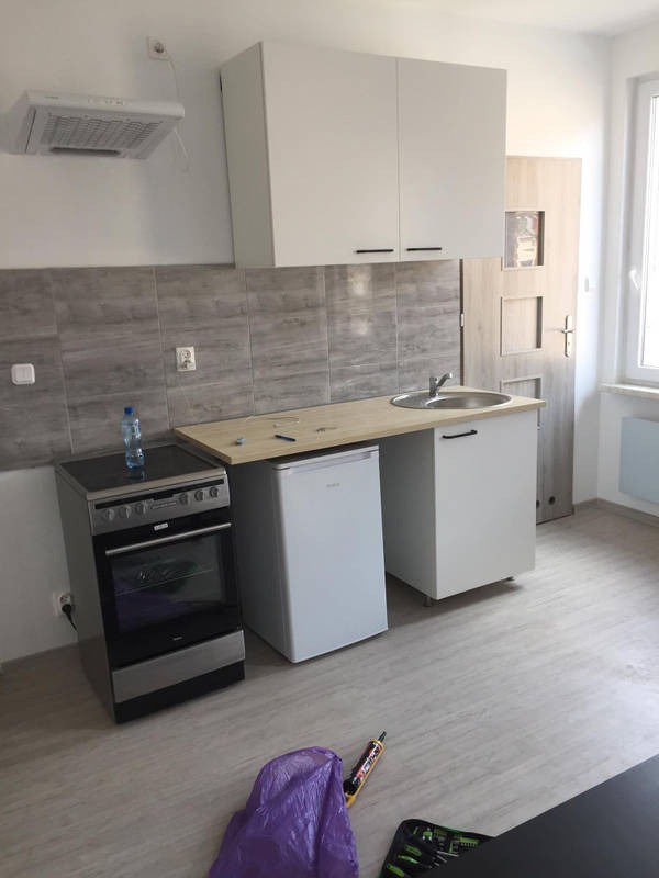 Nowoczesna kuchnia w trakcie montażu: białe szafki wiszące i dolne, kuchenka elektryczna, lodówka pod blatem, widoczne narzędzia i silikon na podłodze.