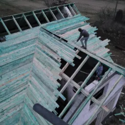 Widok z góry na konstrukcję dachu z zielonych desek, z pracującym na nim dekarzem i fragmentem murowanego domu w tle.
