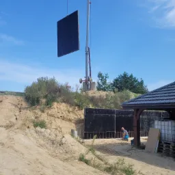 Montaż paneli ściennych na fundamencie zbrojonym, widoczny pracownik przy pracy, żuraw przenoszący panel i teren budowy.