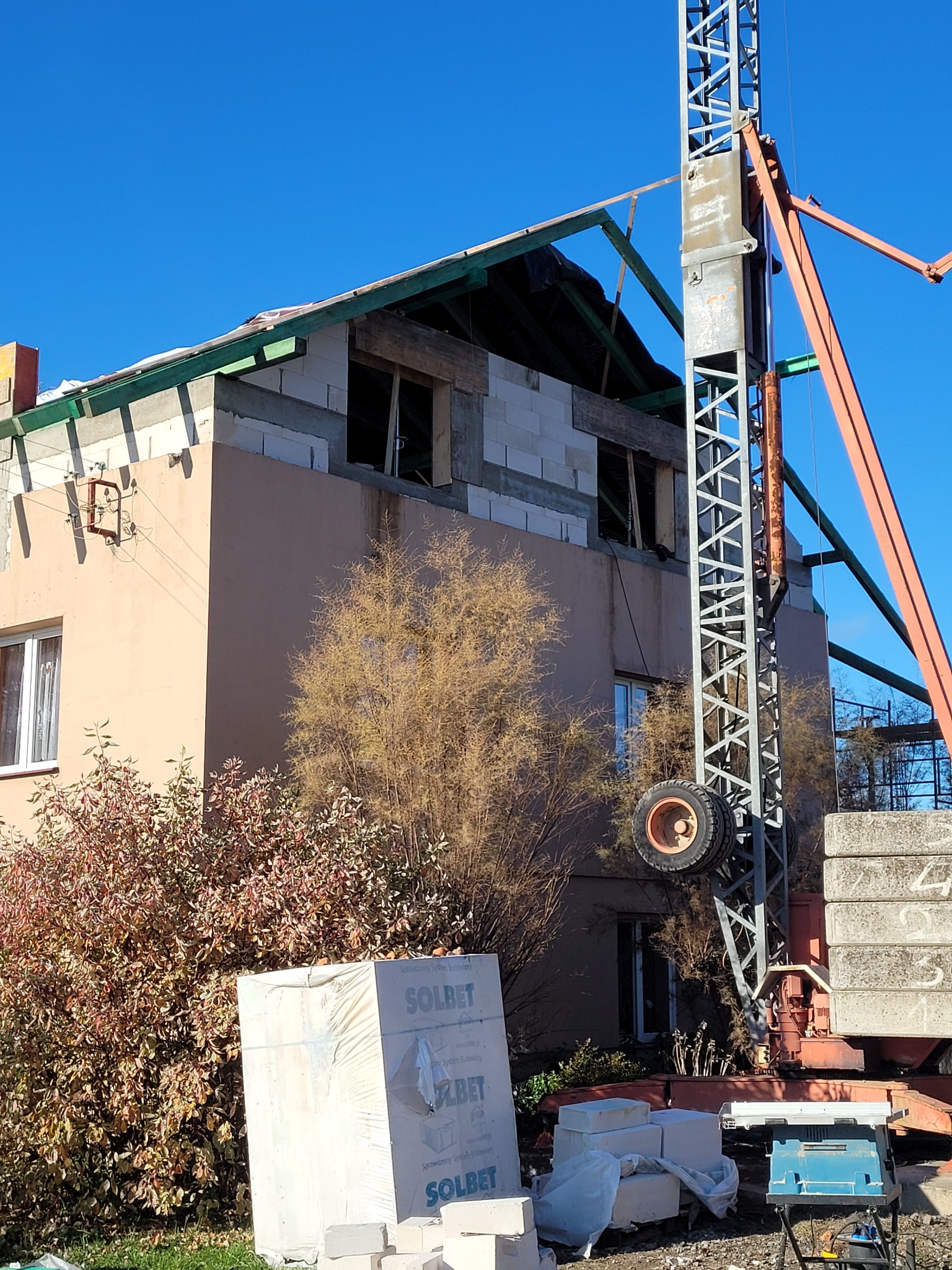 Wznoszenie dwupiętrowego budynku z pustaków, widoczny dźwig budowlany, materiały budowlane Solbet na pierwszym planie, konstrukcja dachu w trakcie realizacji.