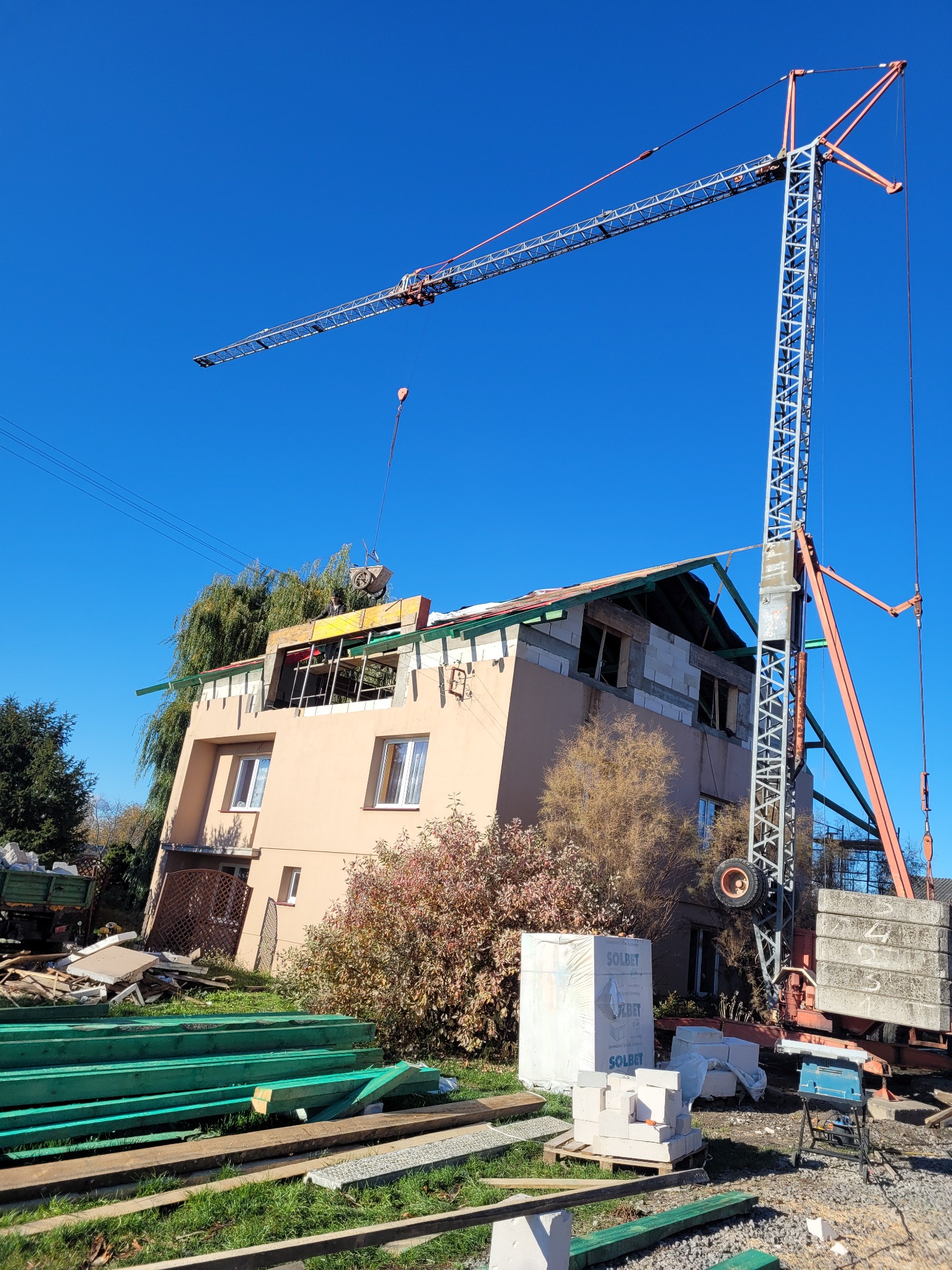 Wznoszenie dwukondygnacyjnego domu z pustaków silikatowych z użyciem dźwigu wieżowego, widoczne elementy konstrukcji dachu, materiały budowlane na placu budowy, słoneczny dzień.