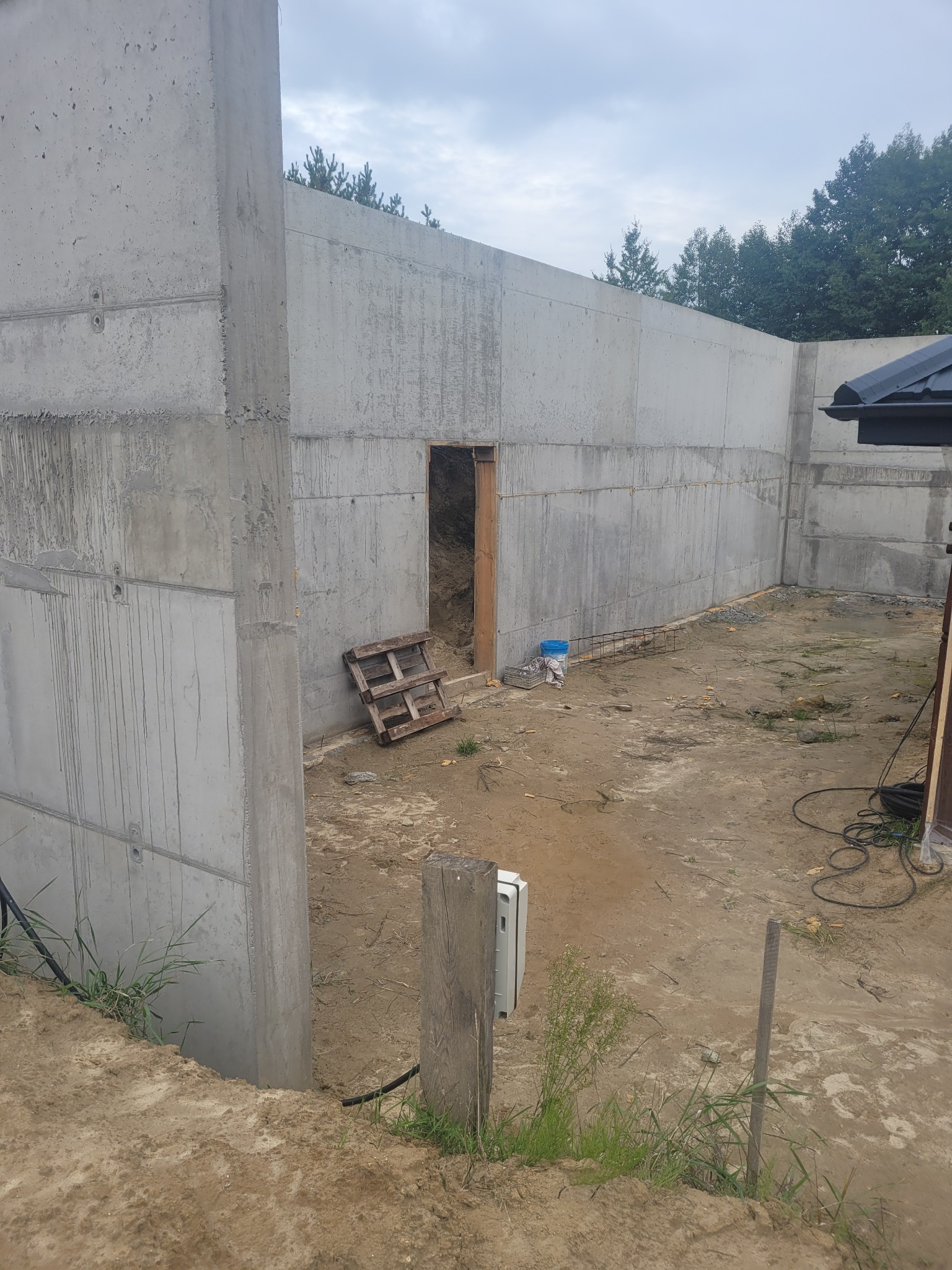 Surowe betonowe ściany w trakcie budowy, widoczne ślady po szalunku, drewniana paleta i niebieskie wiaderko leżące przy ścianie, wycięty otwór drzwiowy w ścianie, teren budowy w trakcie prac.