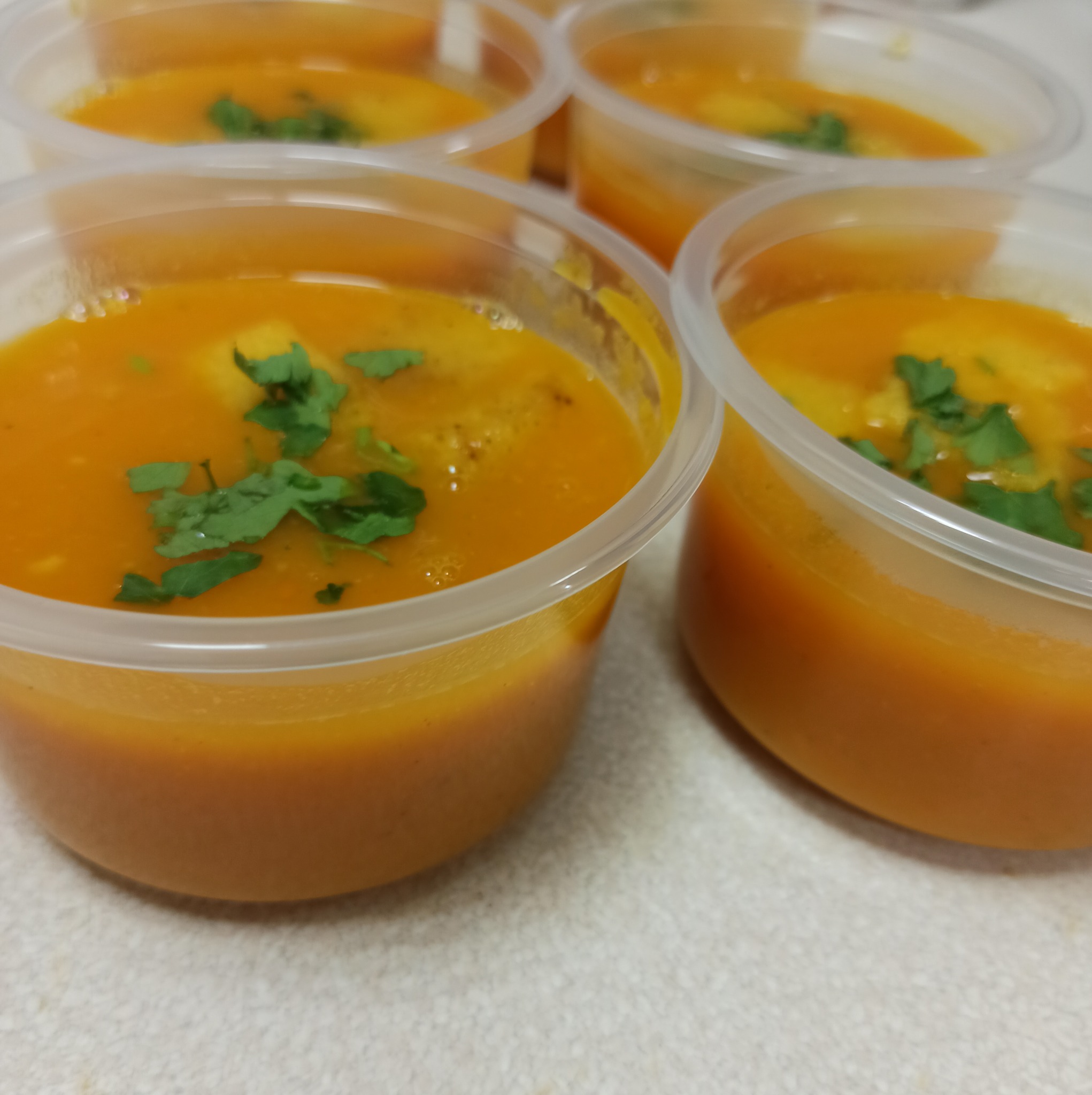Porcje kremowej pomarańczowej zupy dyniowej z zieloną pietruszką w przezroczystych plastikowych pojemnikach, gotowe do dostarczenia.