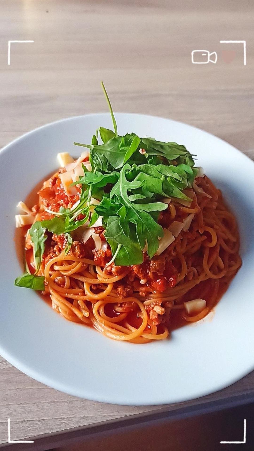 Porcja spaghetti bolognese na białym talerzu, udekorowana rukolą i płatkami sera, sfotografowana z góry na drewnianym stole.
