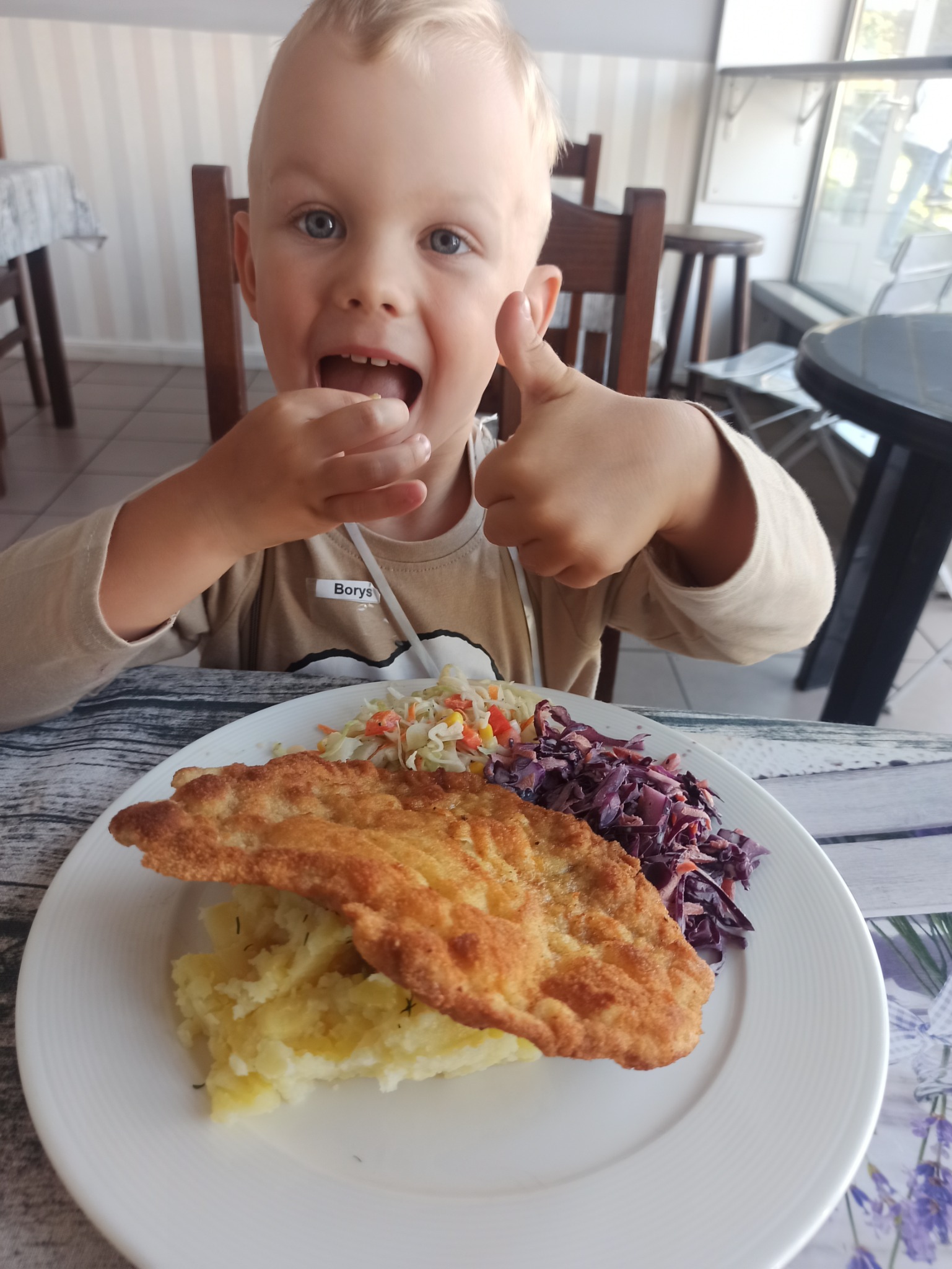 Uśmiechnięty chłopiec z identyfikatorem 'Borys' zajada kotlet schabowy z puree i surówkami, pokazując kciuk w górę.
