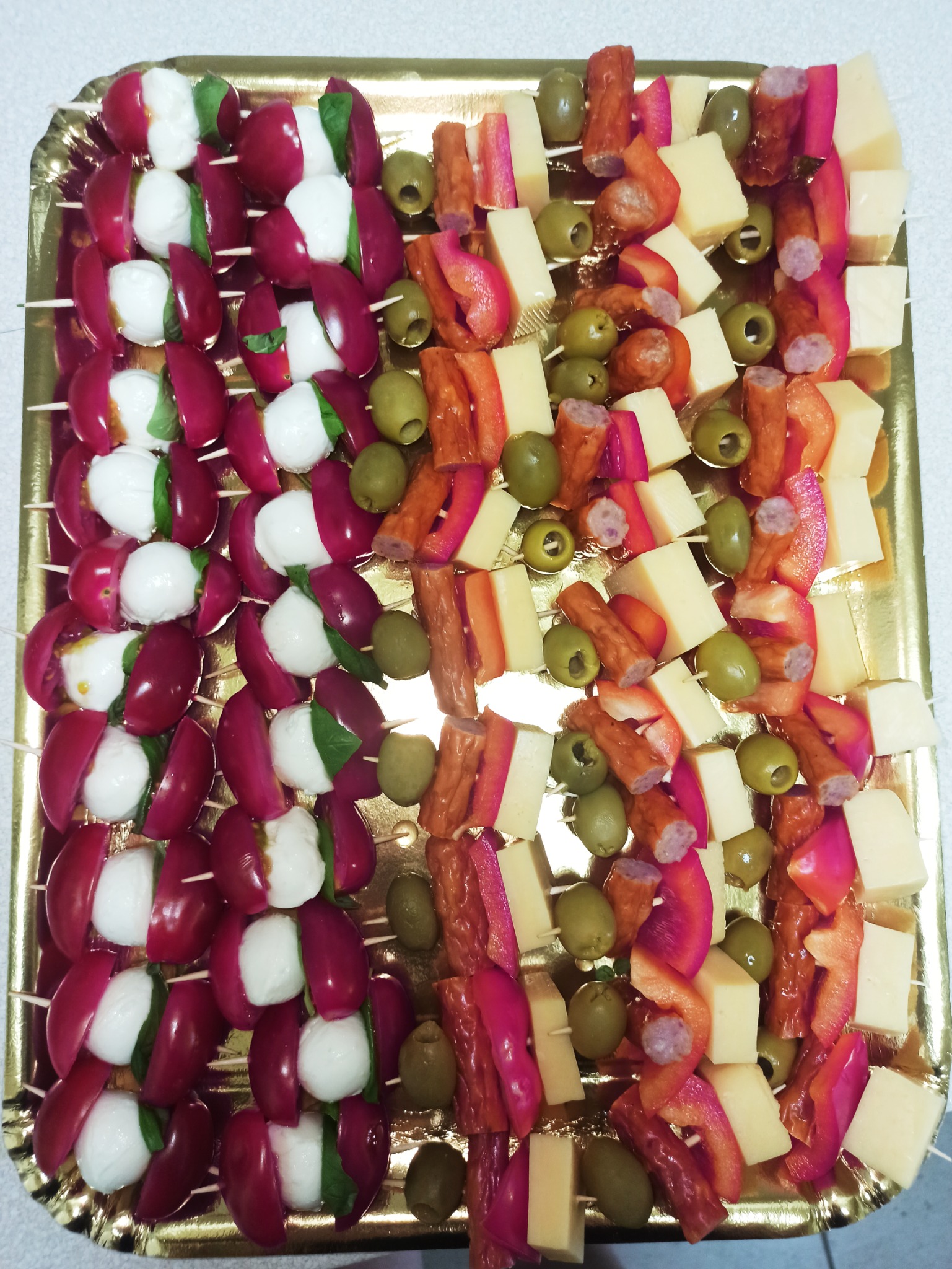 Dekoracyjna złota taca z przekąskami na przyjęcie: szaszłyki Caprese z pomidorków koktajlowych, mozzarelli i bazylii oraz koreczki z sera, oliwek i kawałków wędliny.