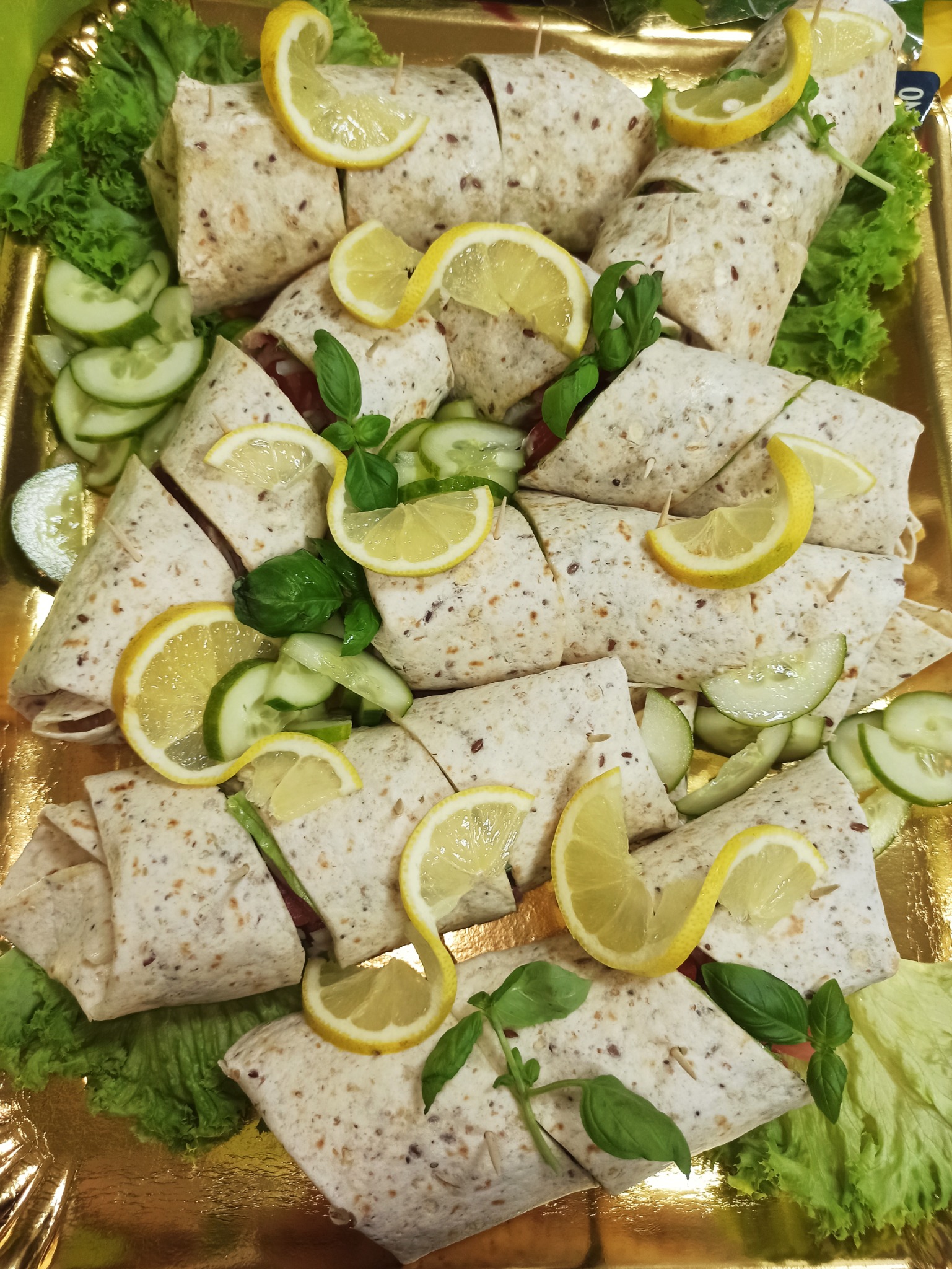 Elegancko ułożone na złotej tacy tortille z nadzieniem, udekorowane plasterkami cytryny, ogórkiem i świeżą bazylią, tworzące apetyczną kompozycję przekąsek.