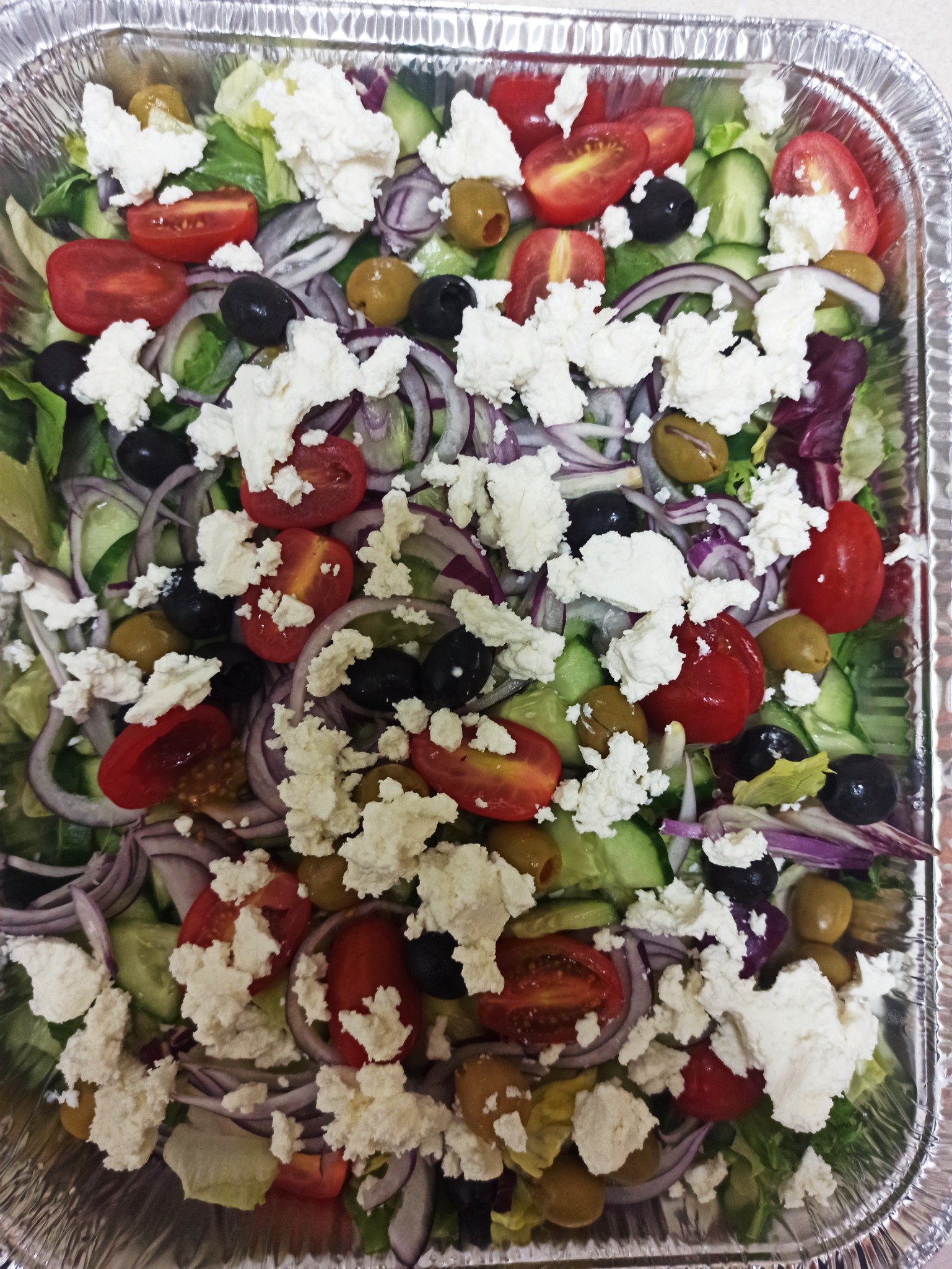 Sałatka grecka z serem feta, pomidorkami koktajlowymi, ogórkiem, czerwoną cebulą i oliwkami, ułożona w aluminiowym pojemniku cateringowym.