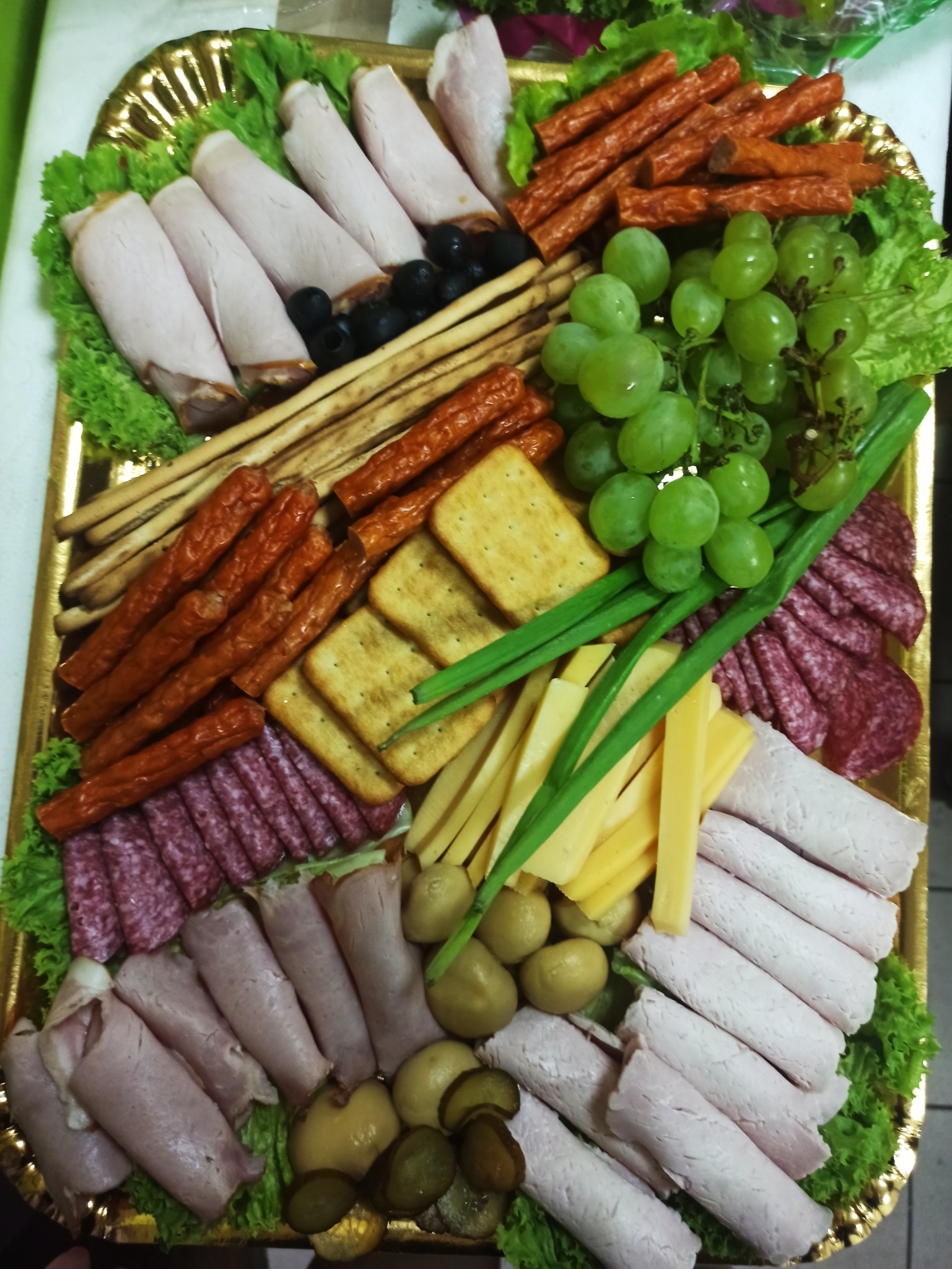 Dekoracyjna złota taca z przekąskami: wędliny zwinięte w rulony, salami w plasterkach, sery w słupkach, krakersy, kabanosy, grissini, zielone winogrona, oliwki, pikle, zielony szczypiorek, ułożone...