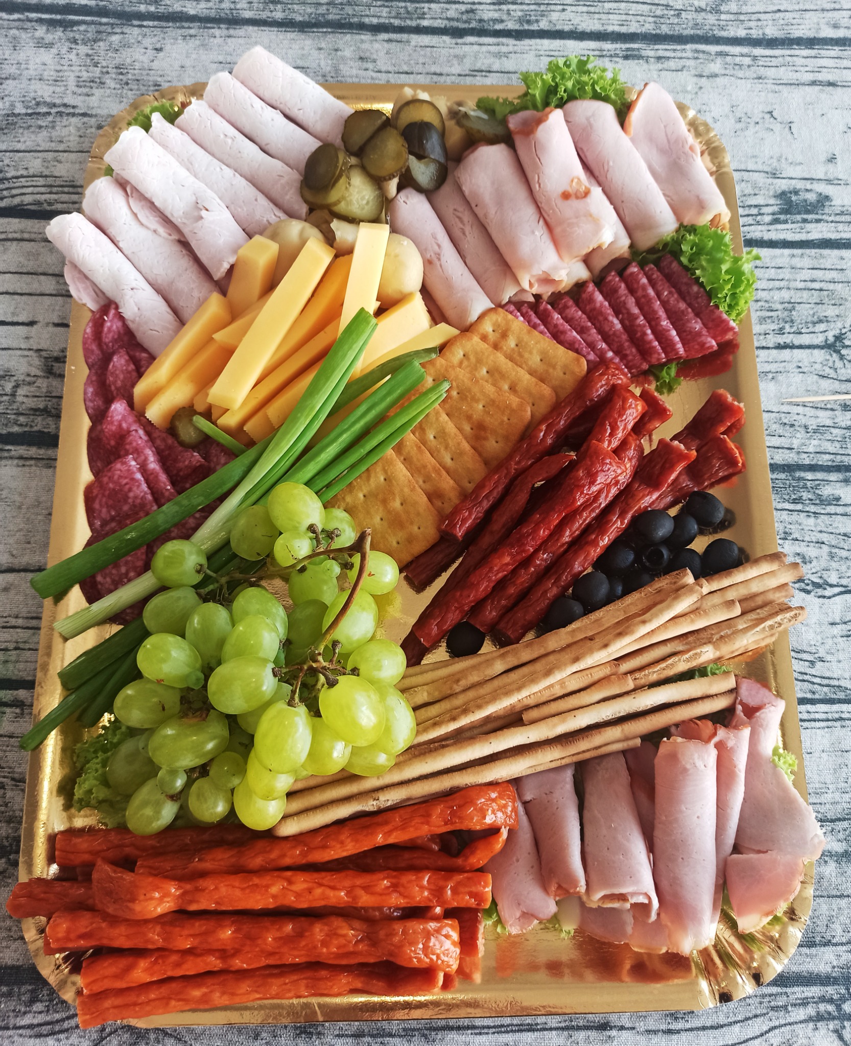 Dekoracyjna złota taca z różnorodnymi przekąskami: wędliny, sery, krakersy, zielone winogrona, ogórki konserwowe, szczypiorek, paluszki grissini i oliwki.