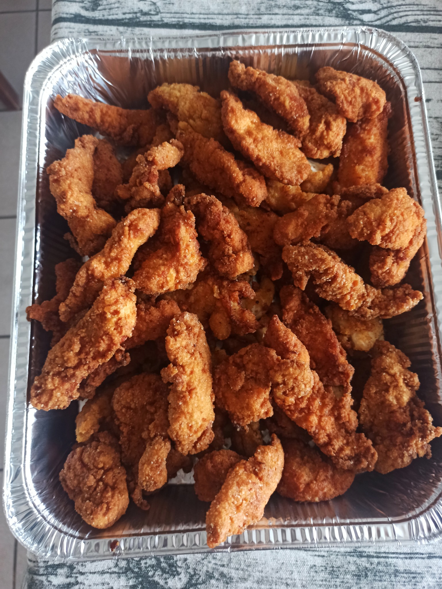 Porcja chrupiących, złocistych kawałków kurczaka w panierce, ułożonych w aluminiowej tacce na tle wzorzystego obrusa.