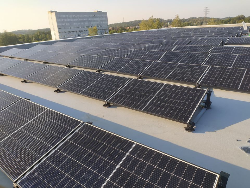 Instalacja fotowoltaiczna na płaskim dachu budynku przemysłowego, z widocznymi systemami montażowymi i budynkiem w tle.