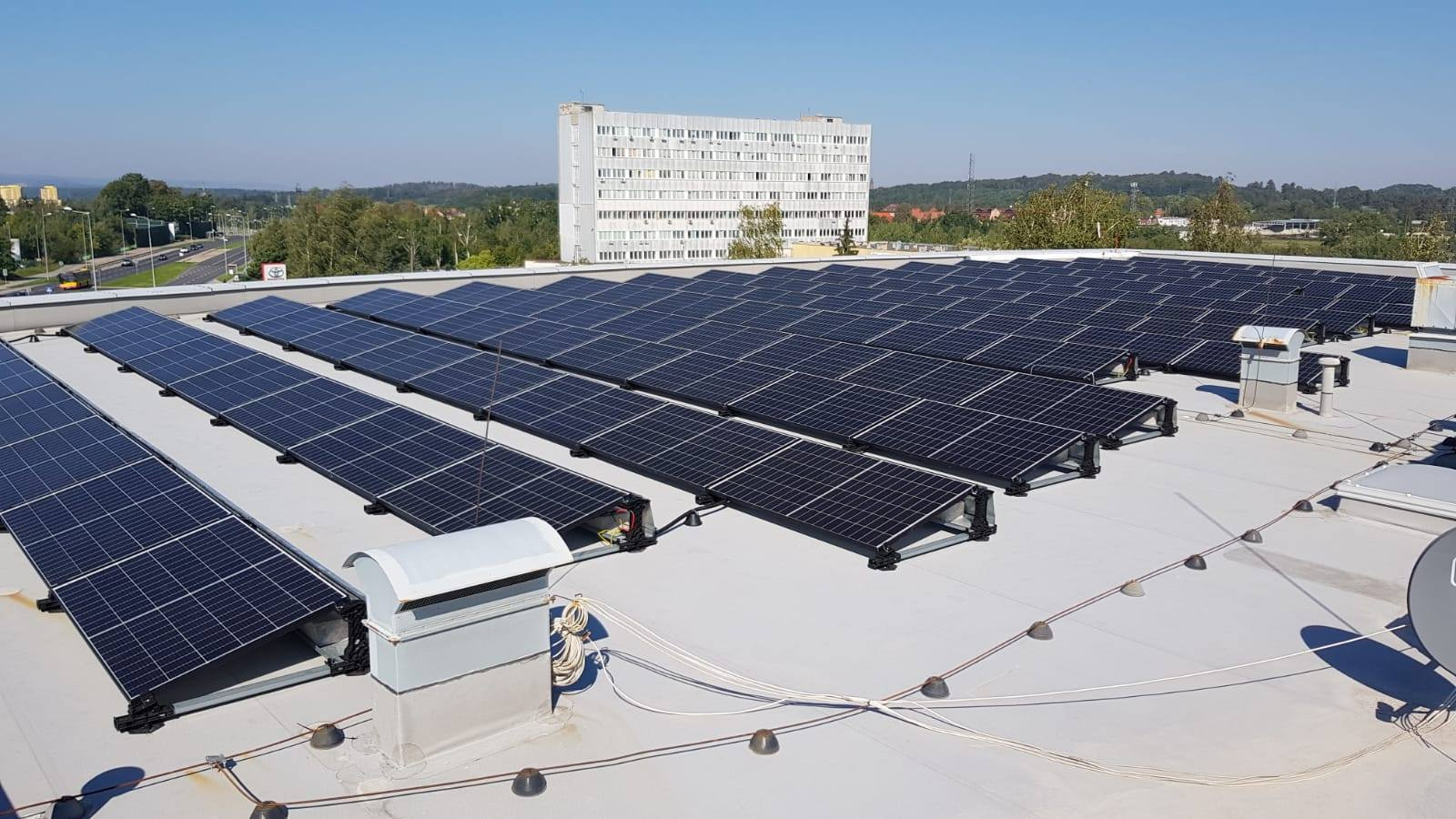 Rzędy paneli słonecznych na płaskim dachu budynku z widokiem na miasto w tle, widoczny komin wentylacyjny i instalacja odgromowa.
