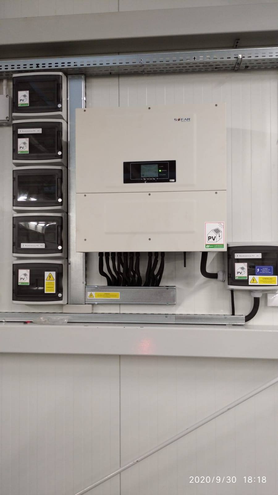 Zamontowany inwerter SOFAR z podłączonymi kablami i zabezpieczeniami w białym pomieszczeniu technicznym. Widoczne naklejki ostrzegawcze i oznaczenia PV.