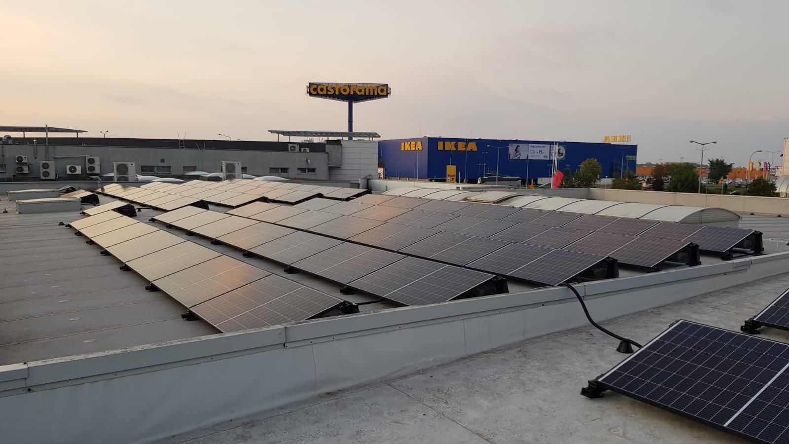 Panele słoneczne na dachu budynku handlowego z widocznymi logotypami Castorama i IKEA w tle, sfotografowane o zmierzchu.