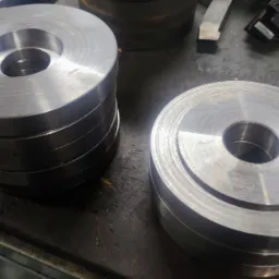 Stos obrobionych, metalowych krążków z otworem, leżących na brudnym stole warsztatowym, obok narzędzi.