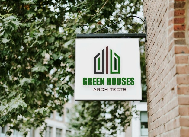 Szyld reklamowy z logo firmy Green Houses Architects, zawieszony na ozdobnym wsporniku przy ścianie z czerwonej cegły, otoczony zielenią drzew.