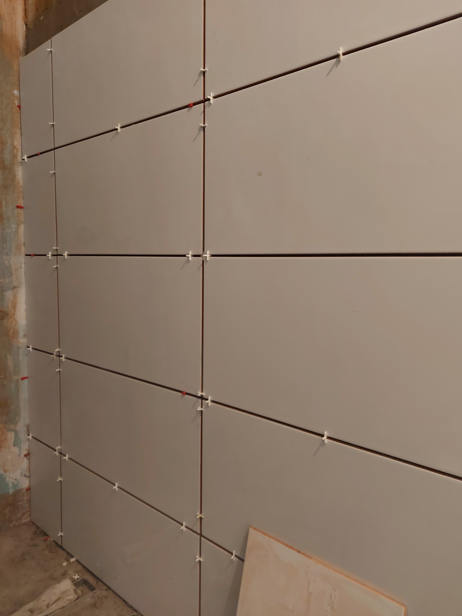 Świeżo ułożone, prostokątne, jasnoszare płytki na ścianie, z widocznymi krzyżykami dystansowymi i klinami poziomującymi, z surowym fragmentem ściany w tle.