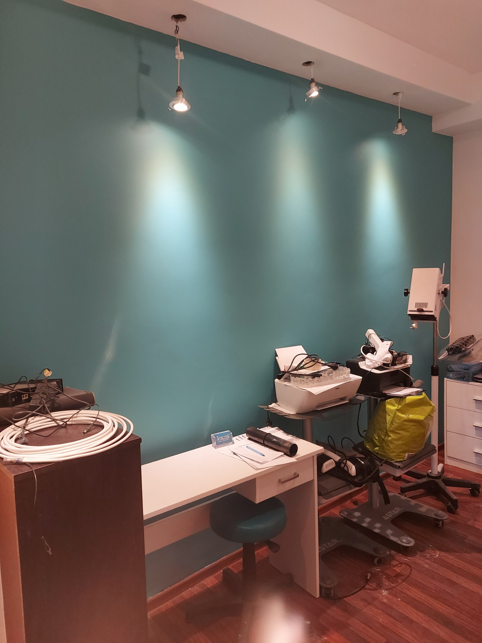 Świeżo pomalowane turkusowe ściany w pomieszczeniu z trzema lampami sufitowymi, widoczne meble biurowe i sprzęt medyczny.