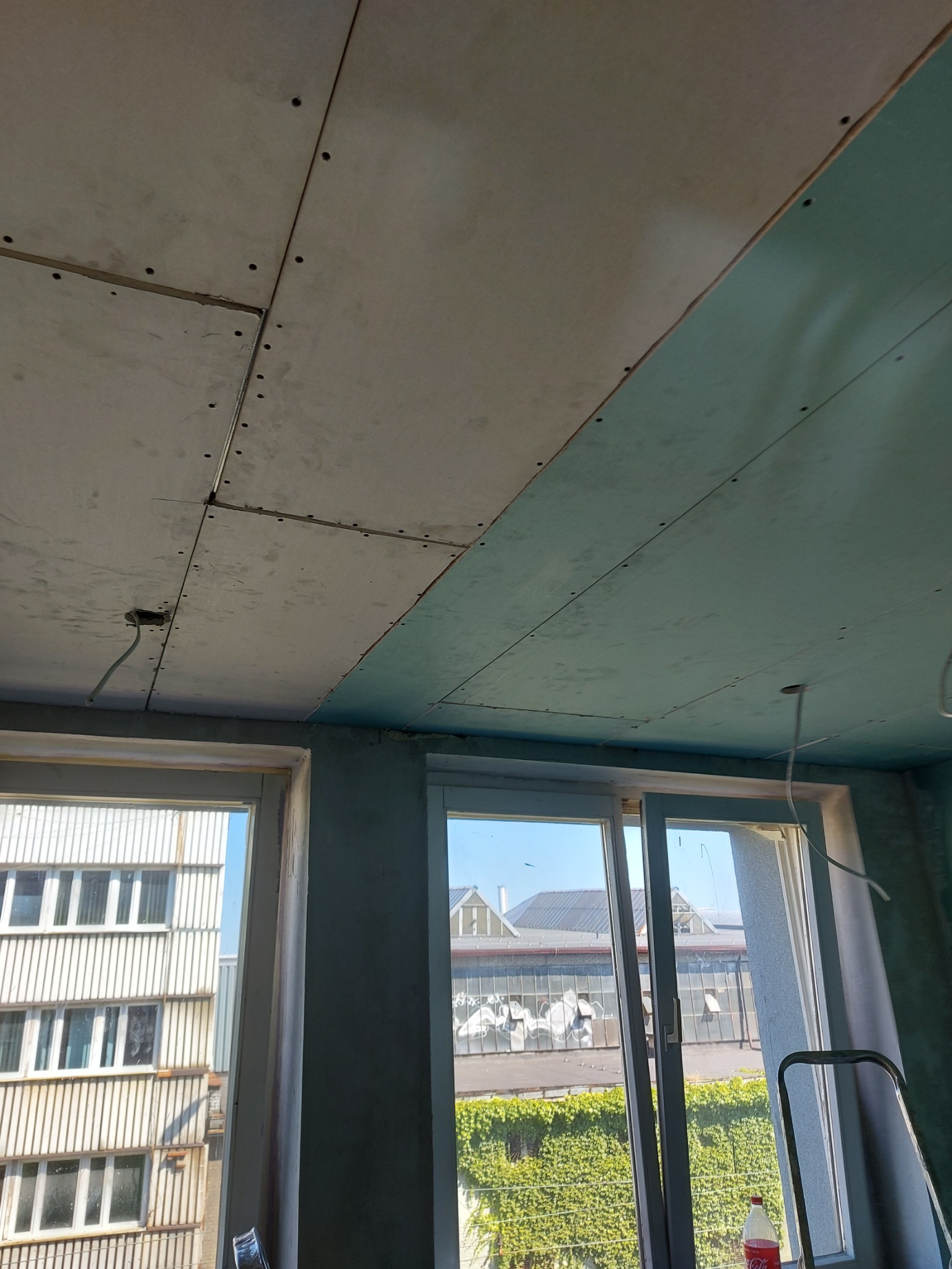 Widok sufitu z zamontowaną płytą kartonowo-gipsową, widoczne łączenia płyt, otwór na instalację elektryczną oraz okna z widokiem na budynek mieszkalny i zieleń.