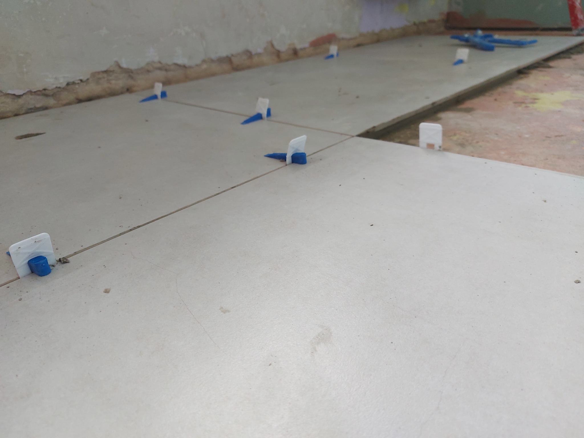 Układanie jasnych, kwadratowych płytek podłogowych z użyciem systemu poziomowania, widoczny surowy stan ściany w tle.