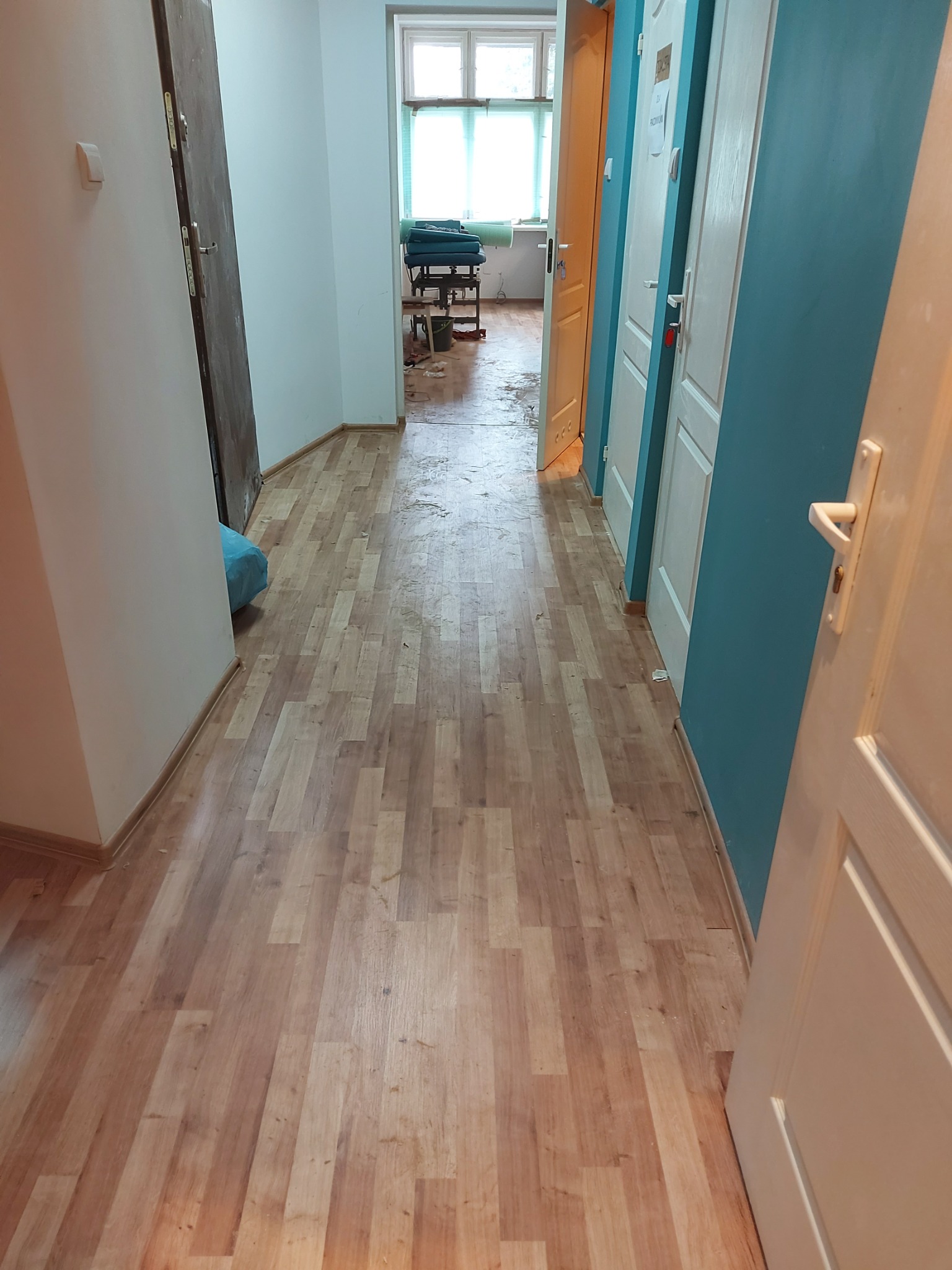 Korytarz z panelami podłogowymi o wzorze imitującym drewno, widoczne ślady po remoncie, niebieska ściana i białe drzwi.