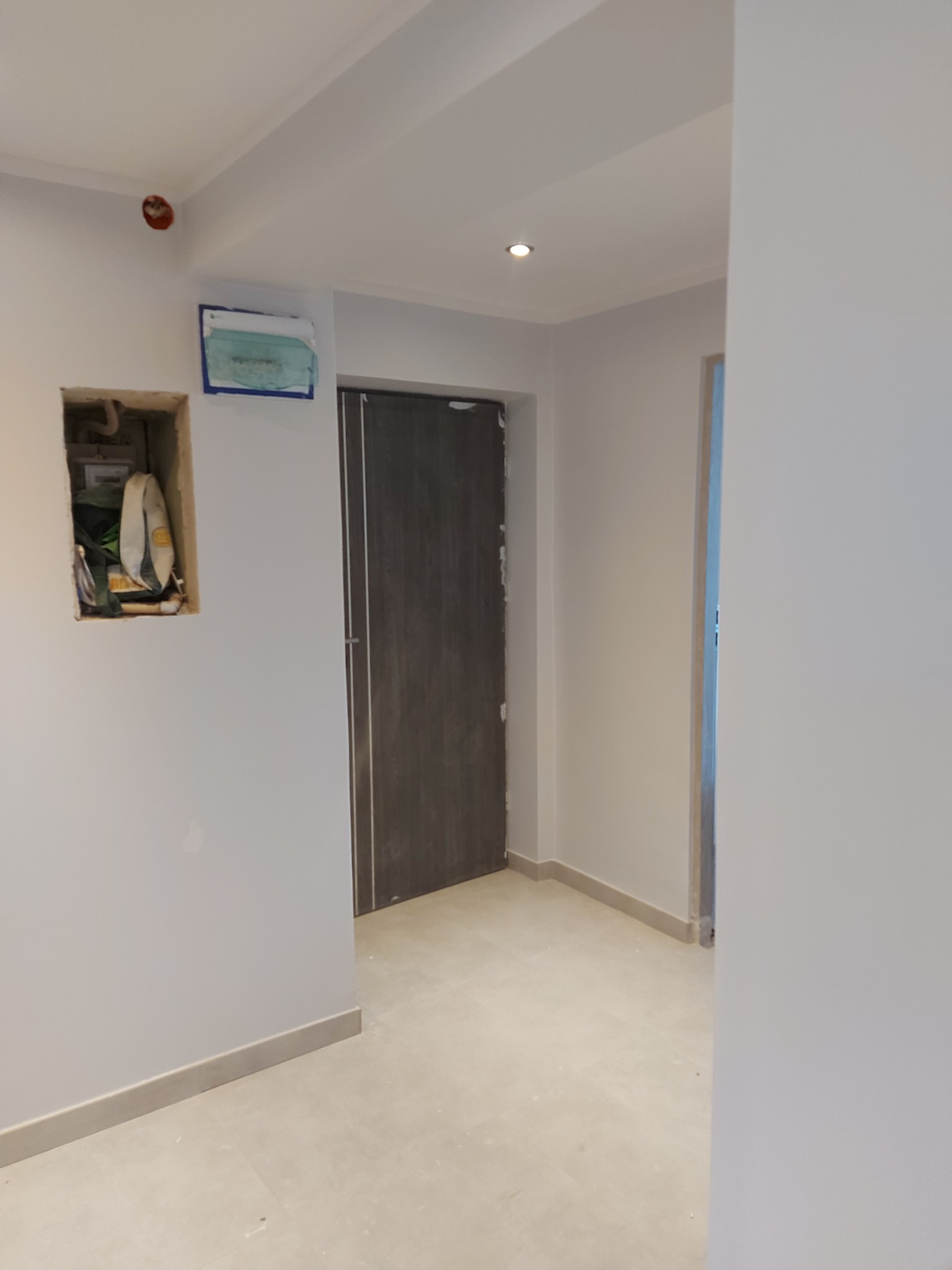 Wnętrze w trakcie prac wykończeniowych: ściany pomalowane na szaro, widoczna niezamaskowana skrzynka elektryczna w ścianie, zabezpieczona folią tablica, szara ościeżnica drzwiowa.