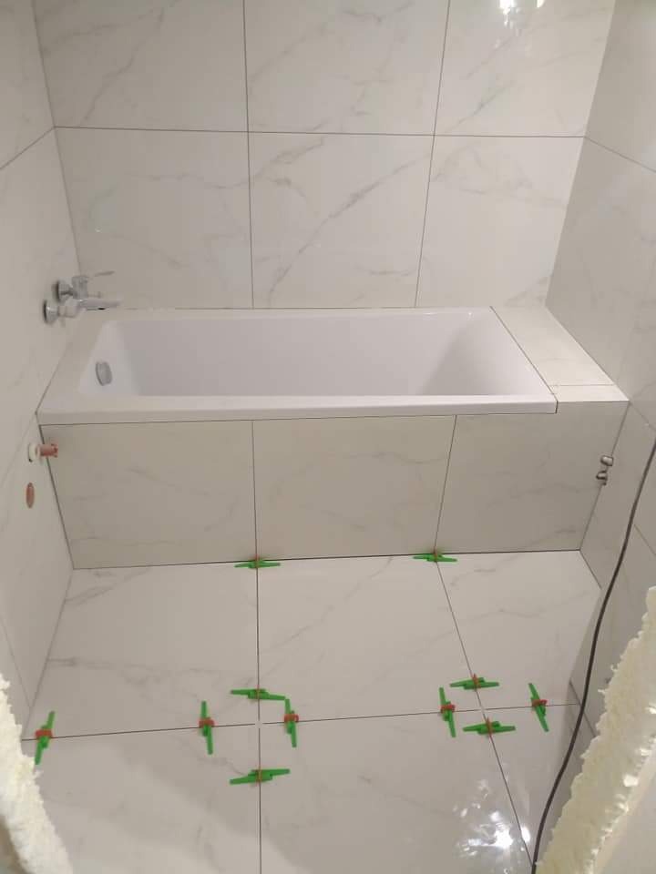 Łazienka w trakcie wykańczania z białą wanną obudowaną płytkami imitującymi marmur, widoczne zielone krzyżyki dystansowe między płytkami na podłodze i ścianach.