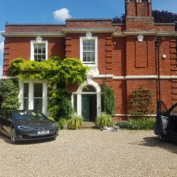 Czerwony, dwupiętrowy dom z cegły z białymi ramami okiennymi i zielonymi pnączami, szary samochód elektryczny i otwarte drzwi innego auta na żwirowym podjeździe w słoneczny dzień.