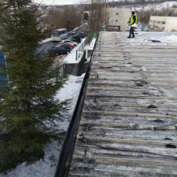 Pracownik w żółtej kamizelce stoi na płaskim, metalowym dachu pokrytym resztkami śniegu i systemem przeciwoblodzeniowym, widok z góry na ulicę z zaparkowanymi samochodami i budynkami w tle.