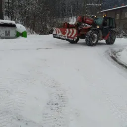 Czerwono-biały pług śnieżny zamontowany na podnośniku teleskopowym usuwa śnieg z drogi, widoczne ślady opon na śniegu, w tle drzewa i budynek.