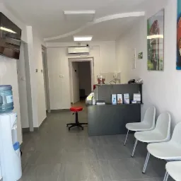 Wnętrze poczekalni gabinetu medycznego: szare płytki podłogowe, białe ściany, rząd białych krzeseł, dystrybutor wody, czerwony taboret i recepcja z ulotkami informacyjnymi dla pacjentów.