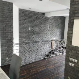 Ściana wykończona szarą cegłą dekoracyjną w Rabce-Zdroju, widoczne ozdobne metalowe balustrady schodów i ciemna drewniana podłoga, oświetlenie punktowe.