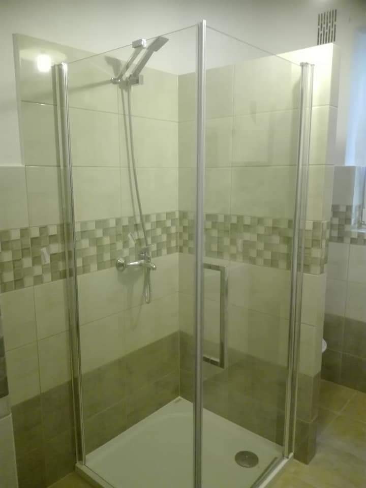 Nowoczesna kabina prysznicowa z chromowanymi profilami, jasnymi płytkami i dekoracyjnym pasem mozaiki, widoczna armatura i odpływ liniowy.