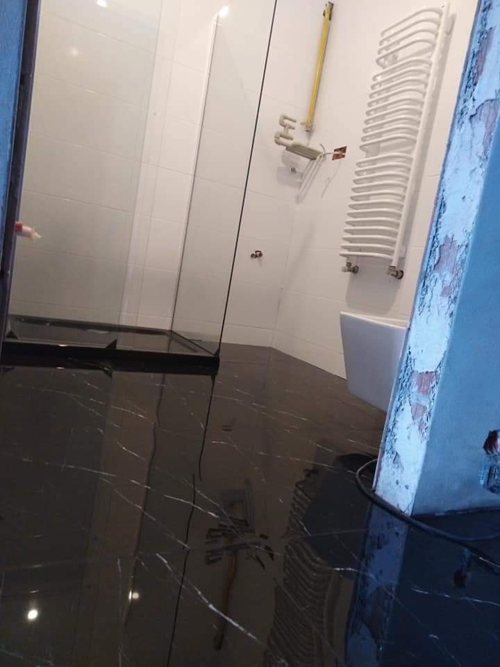 Błyszcząca, czarna marmurowa posadzka w łazience z kabiną prysznicową i grzejnikiem drabinkowym, widoczne ślady niedokończonych prac wykończeniowych na ościeżnicy.