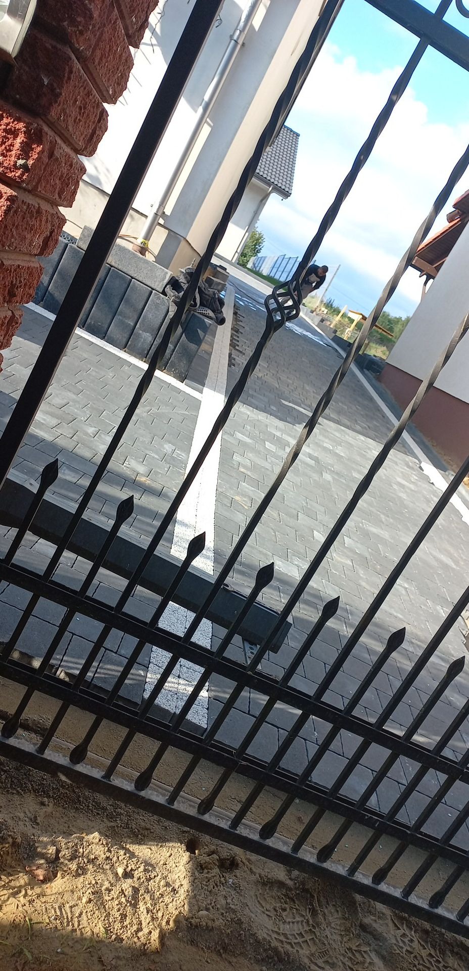 Szara kostka brukowa ułożona wzdłuż białego pasa, widoczna przez kute, metalowe ogrodzenie z dekoracyjnymi elementami.