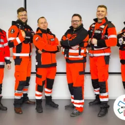 Grupa sześciu ratowników medycznych w pomarańczowych uniformach z naszywkami, stojących w rzędzie na tle białej ściany, z logo MIMED w prawym dolnym rogu.